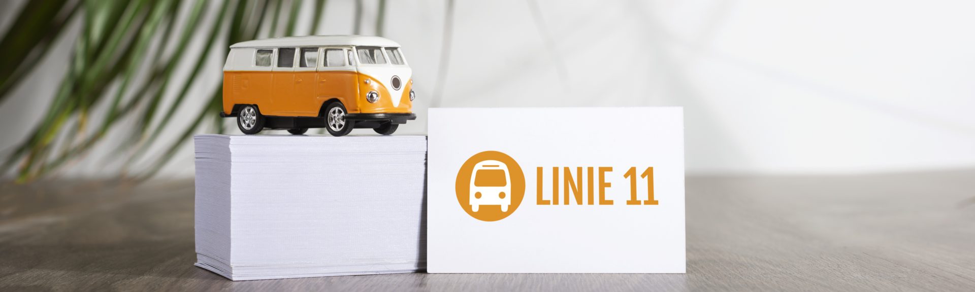 Auf einem Holztisch steht ein Stabel mit Visitenkarten. Auf dem Stapel befindet sich ein orangefarbener Spielzeug-VW-Bus mit weißem Dach. An den Stapel angelehnt steht eine einzelne Visitenkarte mit dem Logo der Linie 11.