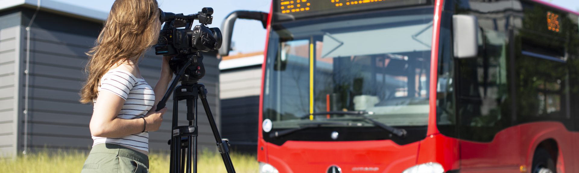 In der rechten Bildhälfte steht ein roter Bus aus dem Nahverkehr. Links steht sich eine junge Fraz mit langen braunen Haaren und sommerlicher Kleidung. Sie schaut durch den Sucher einer Kamera, die auf einem Stativ steht und zum Bus zeigt.
