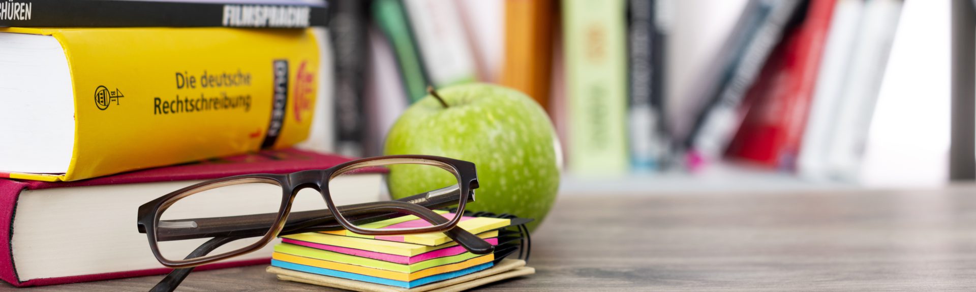 Auf einem Holztisch, der vor einem Bücherregal steht, befinden sich links drei weitere Bücher. Davor befinden sich ein Stapel Haftnotizzettel, eine zusammengeklappte Brille und ein grüner Apfel.